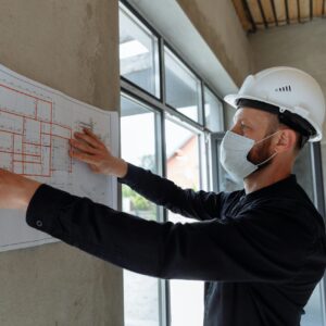 Architect wearing hard hat and mask reviews floor plans on site for new construction project.